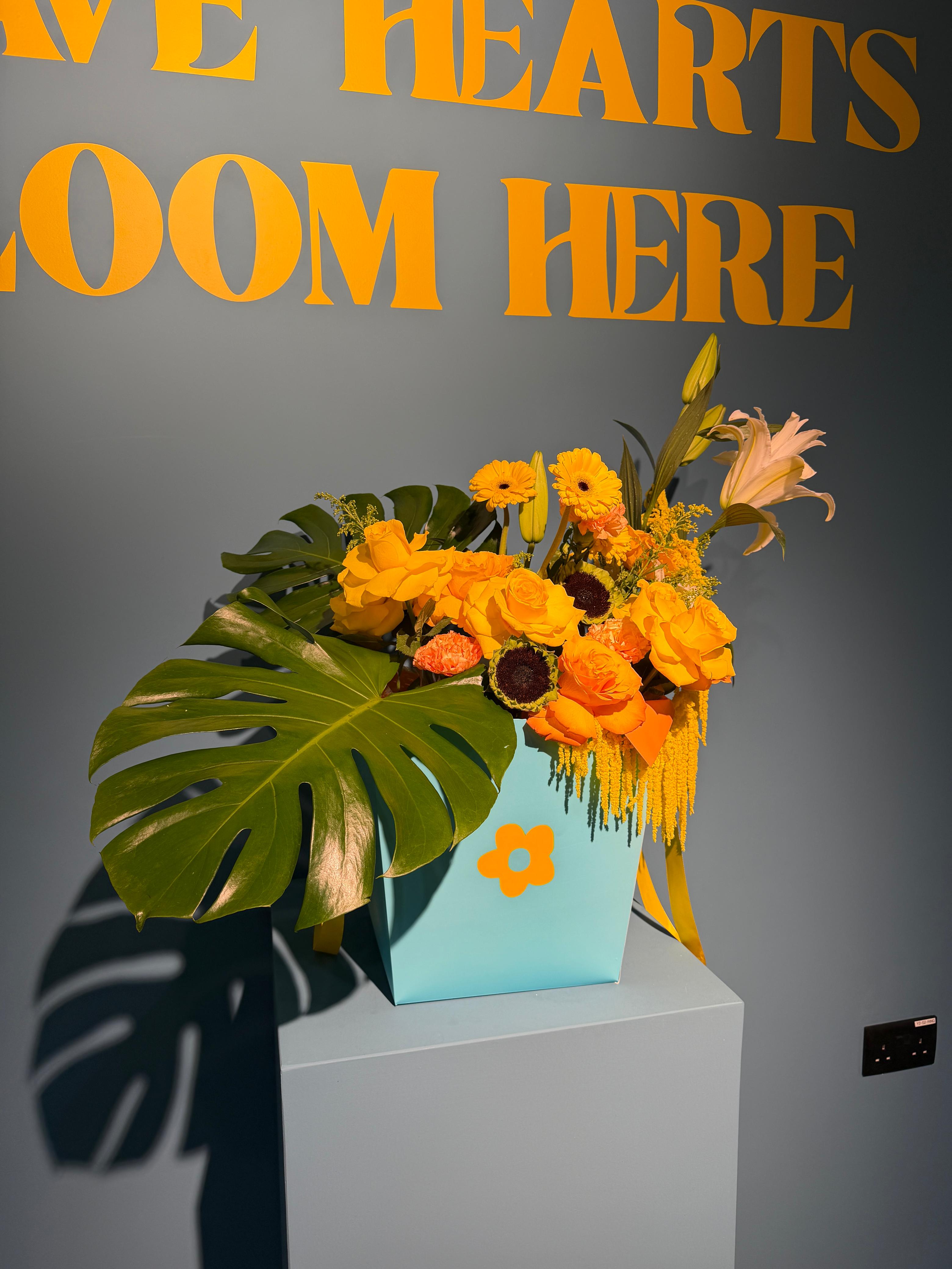 Tropical Sunrise signature box arrangement with yellow and orange roses, sunflowers, and tropical monstera leaves