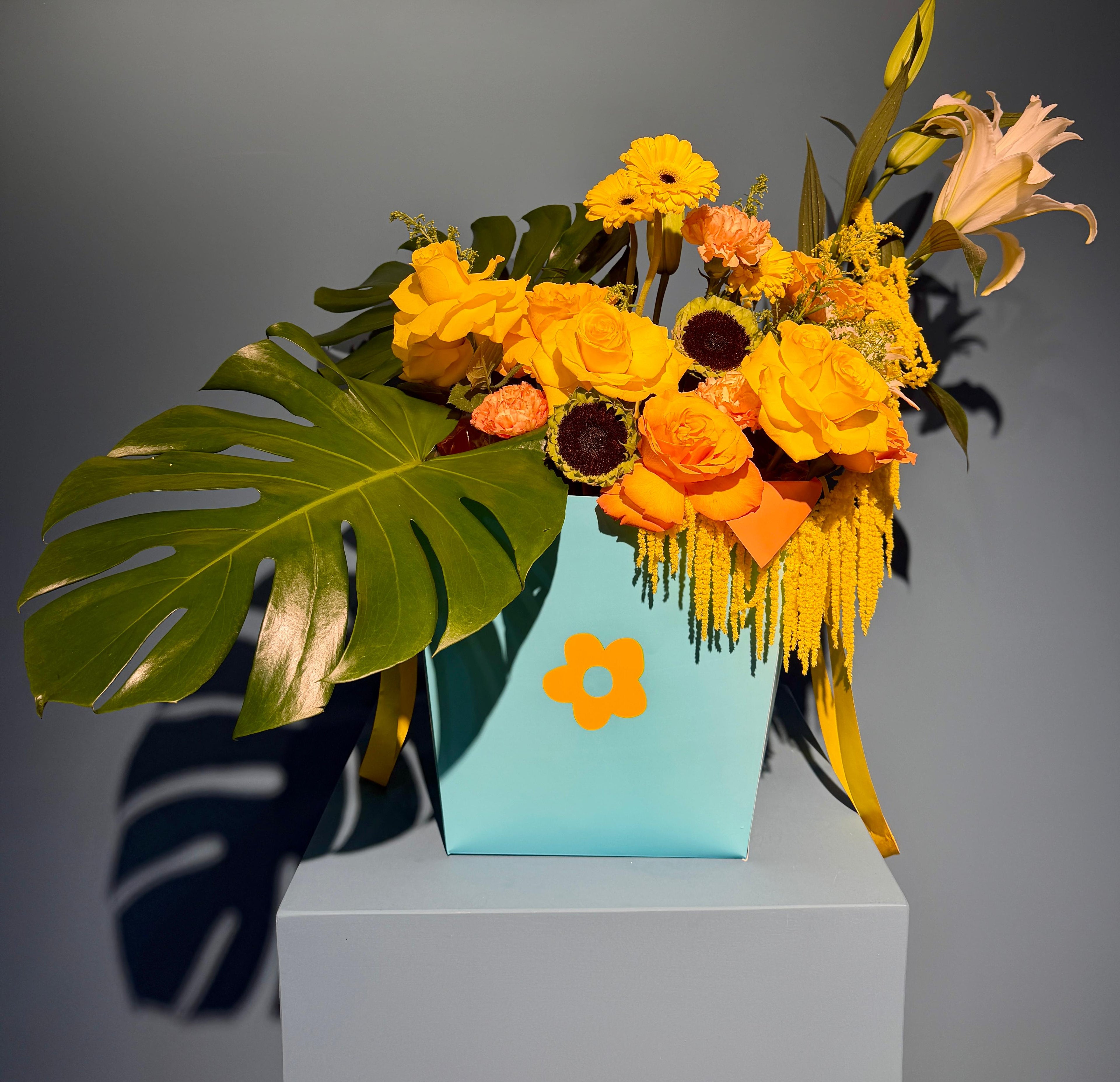 Tropical Sunrise signature box arrangement with yellow and orange roses, sunflowers, and tropical monstera leaves